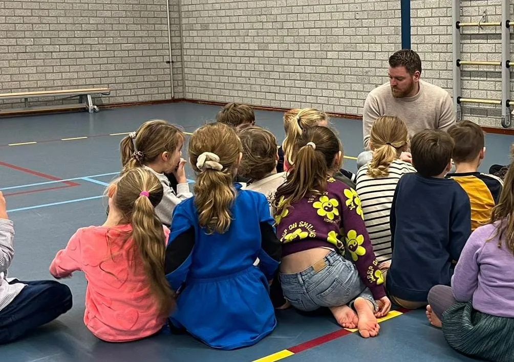Jonge kinderen zitten in de gymzaal op de grond rond een begeleider