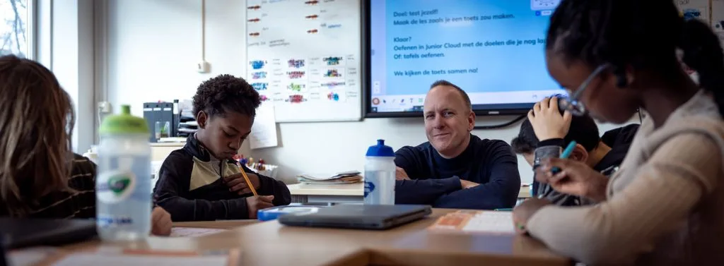 Begeleider zit lachend aan tafel met leerlingen in de klas