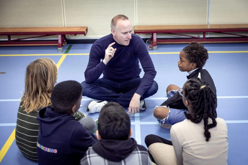 Begeleider zit in de gymzaal in een kring met leerlingen en geeft uitleg